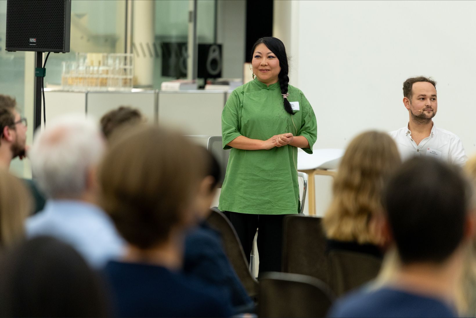Dr. Ning Wang (UZH) speaking at the 20th anniversary of Swissnex, 26.10.2023; feminine-appearing person wearing a green top, black pants and a side braid placed over the left shoulder stands in front of a group of people and gives a lecture